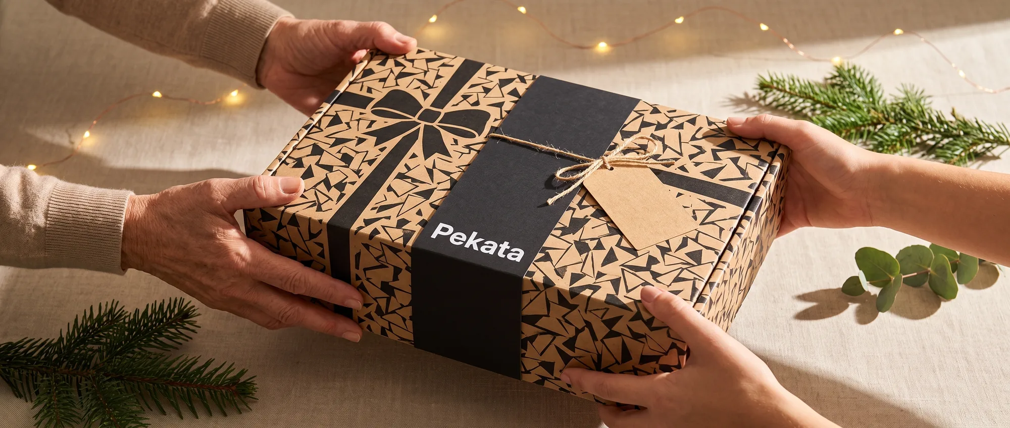 Zwei Paar Hände überreichen und empfangen eine Pekata-Geschenkbox mit Kraftpapieretikett und Trencadís-Muster